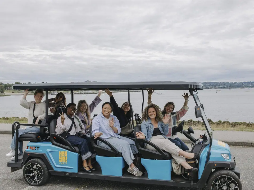 Eight friends waving from a blue HeyYa eight-seat electric cart beside the Vancouver waterfront